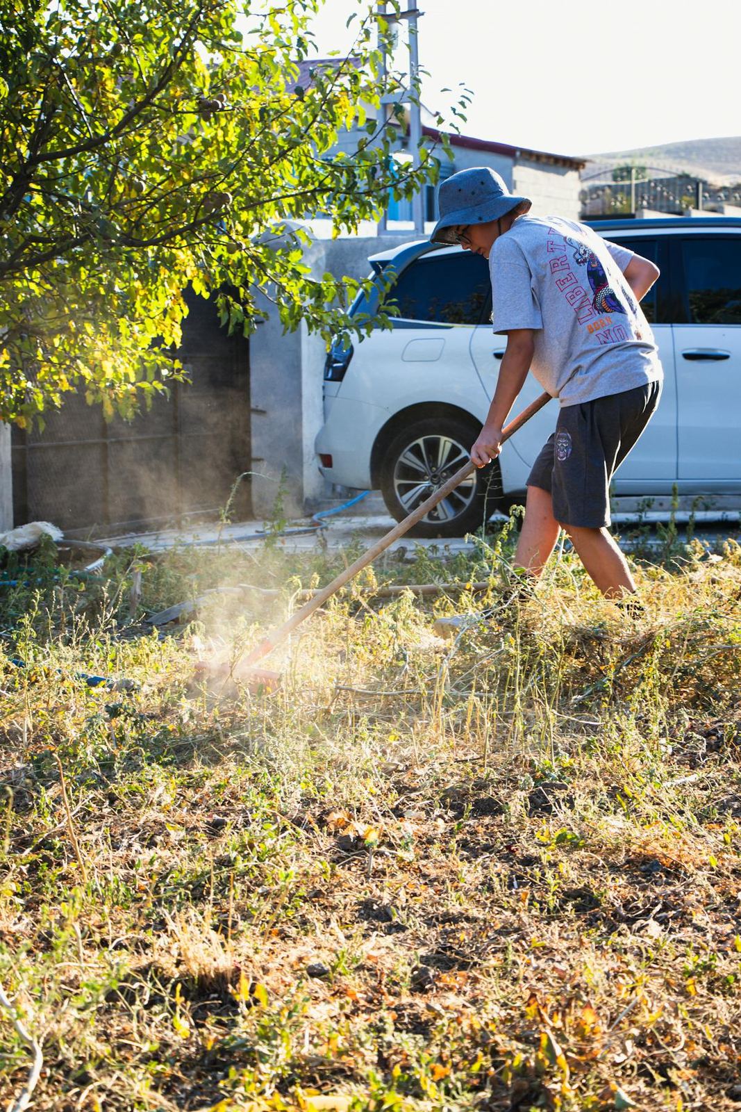 A man in a bucket hat raking leaves in a yard.